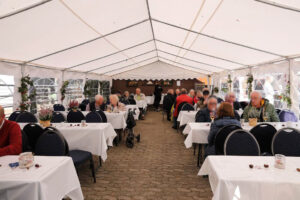 Menschen sitzen an Tischen in einem Zelt, bedeckt mit weißen Tischdecken, umgeben von festlicher Atmosphäre.